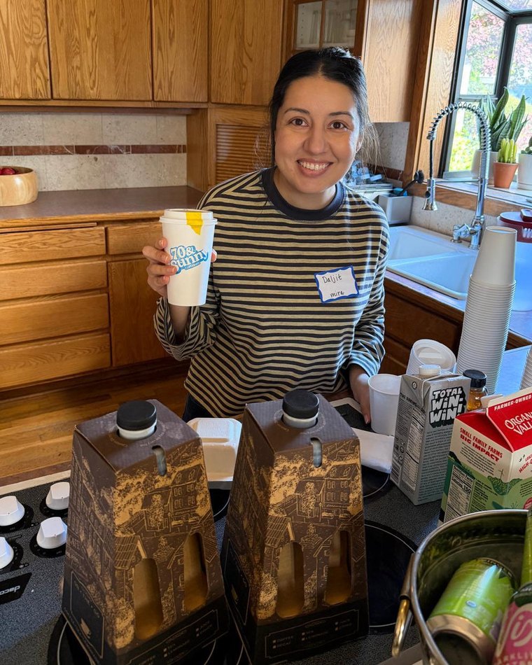 Daljit behind the drinks table in the kitchen, holding a cup, with two urns of coffee, milk and sparkling water set out beside her.
